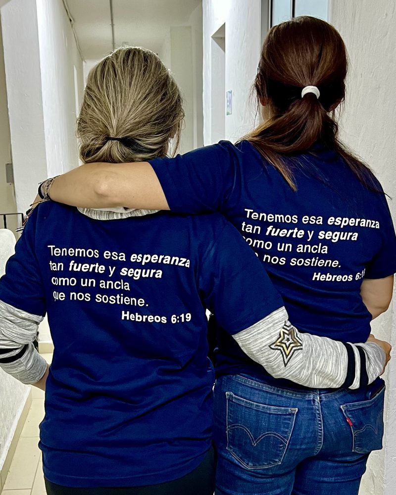 Two women in TIKVA shirts printed with the Hebrews 6:19 verse in Spanish, walking arm in arm in Medellín
